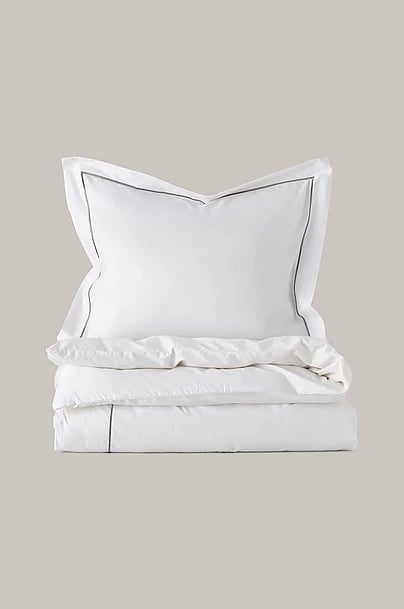 Stack of white bedding including pillow sham, flat sheet, and fitted sheet with gray piping trim on neutral background