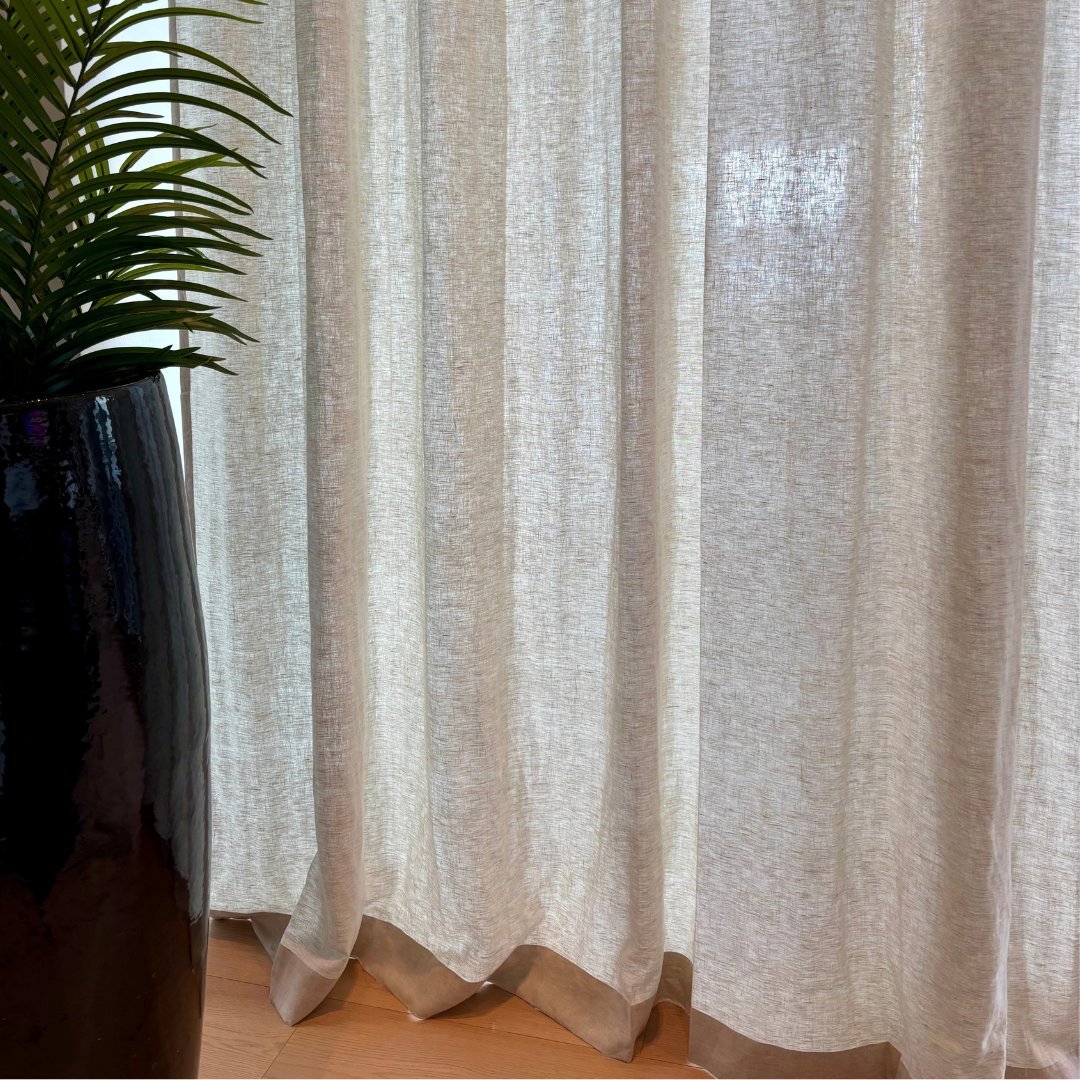 Beige linen curtains hanging against a wall with a black vase containing green foliage on wooden floor